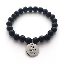 A black beaded bracelet with a round silver charm saying "be here now".