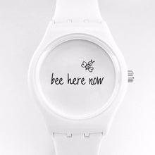 White watch with "bee here now" and a tiny drawing of a bee on the face of the watch.